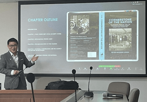 A man in a suit presents in front of a screen that shows the jacket for a book titled, "Cornerstone of the Nation"