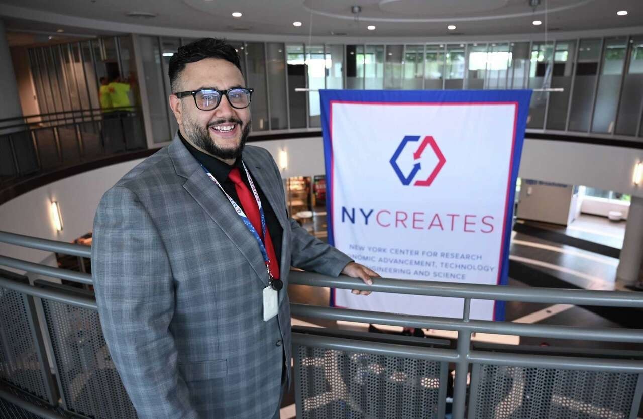 A man wearing a gray checkered suit stands in front of a sign that says "NY CREATES" on it overlooking a circular rotunda indoors.