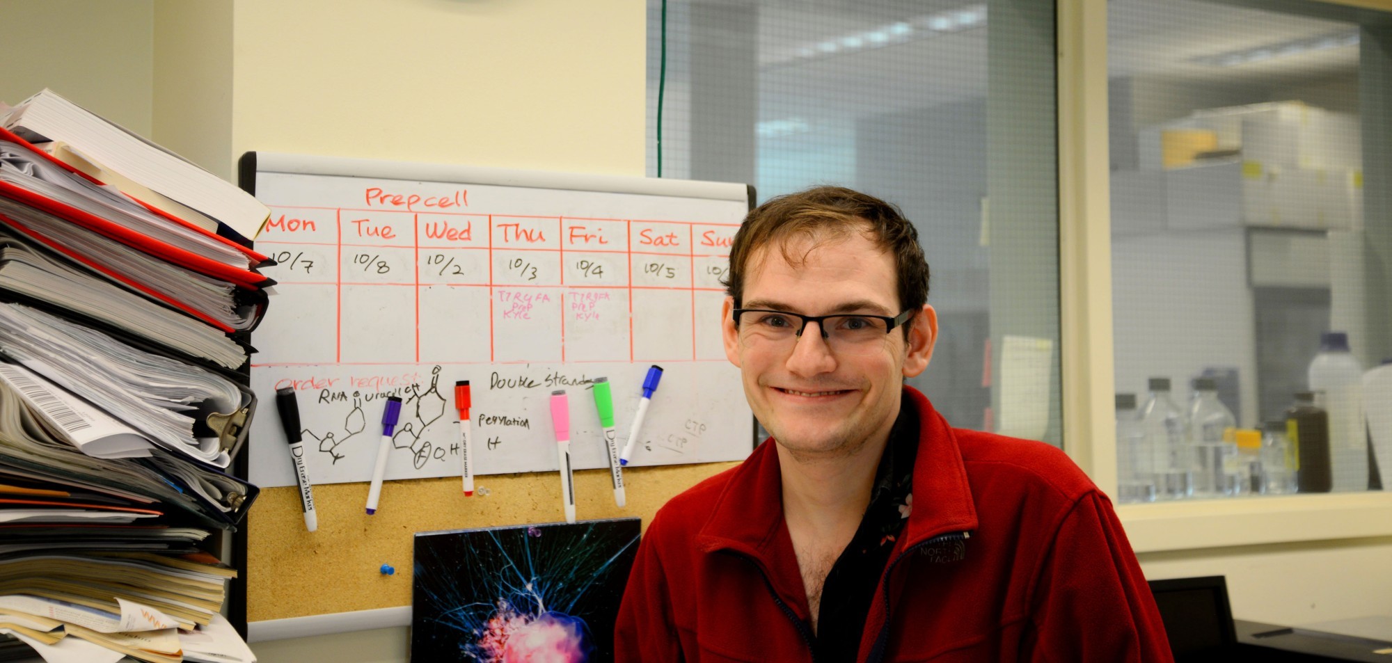 A Niu Research Laboratory team member smiles and poses for a photo while seated at a desk inside the lab. The white board behind him shows a schedule for prepping cells.