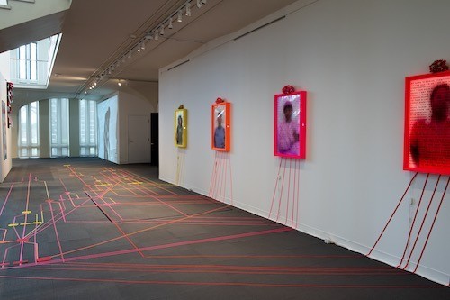 Flourescent gaffers tape create a path on the University Art Museum floor. Several pieces of tape travel up a white wall and lead to three lightbox portraits of individuals covered in text.