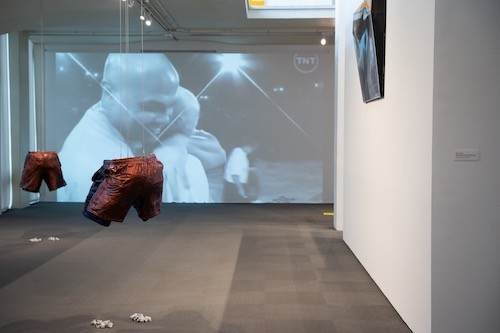 Basketball players hug on a large video screen inside the University Art Museum. Two sculptures of basketball shorts stand nearby.