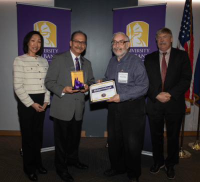 Four people stand in front of UAlbany banners, one holding a purple great Dane bust and one holding a certificate with his name, Richard Albagli, printed on it.
