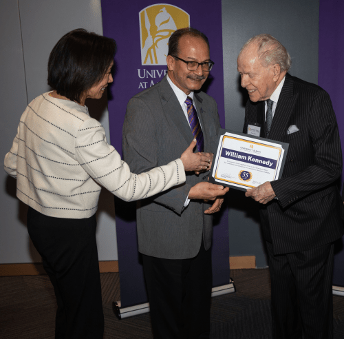 William Kennedy holds a certificate with his name printed on it, while being congratulated by Provost Carol Kim and President Havidán Rodríguez.