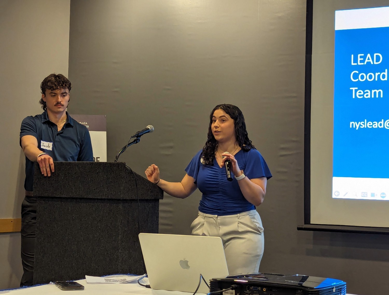 Jacob Schoff and Alexis stand at a dark wooden podium. Alexis is speaking at the mic, wearing a blue top and white pants. A PowerPoint slide at left says 'LEAD Coordinating team'.