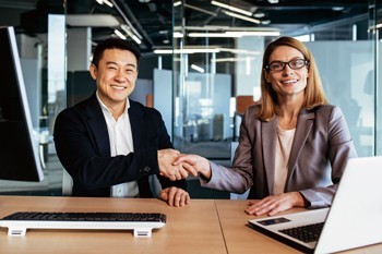 Two professionals shaking hands and smiling while seated at a desk with a keyboard and laptop.