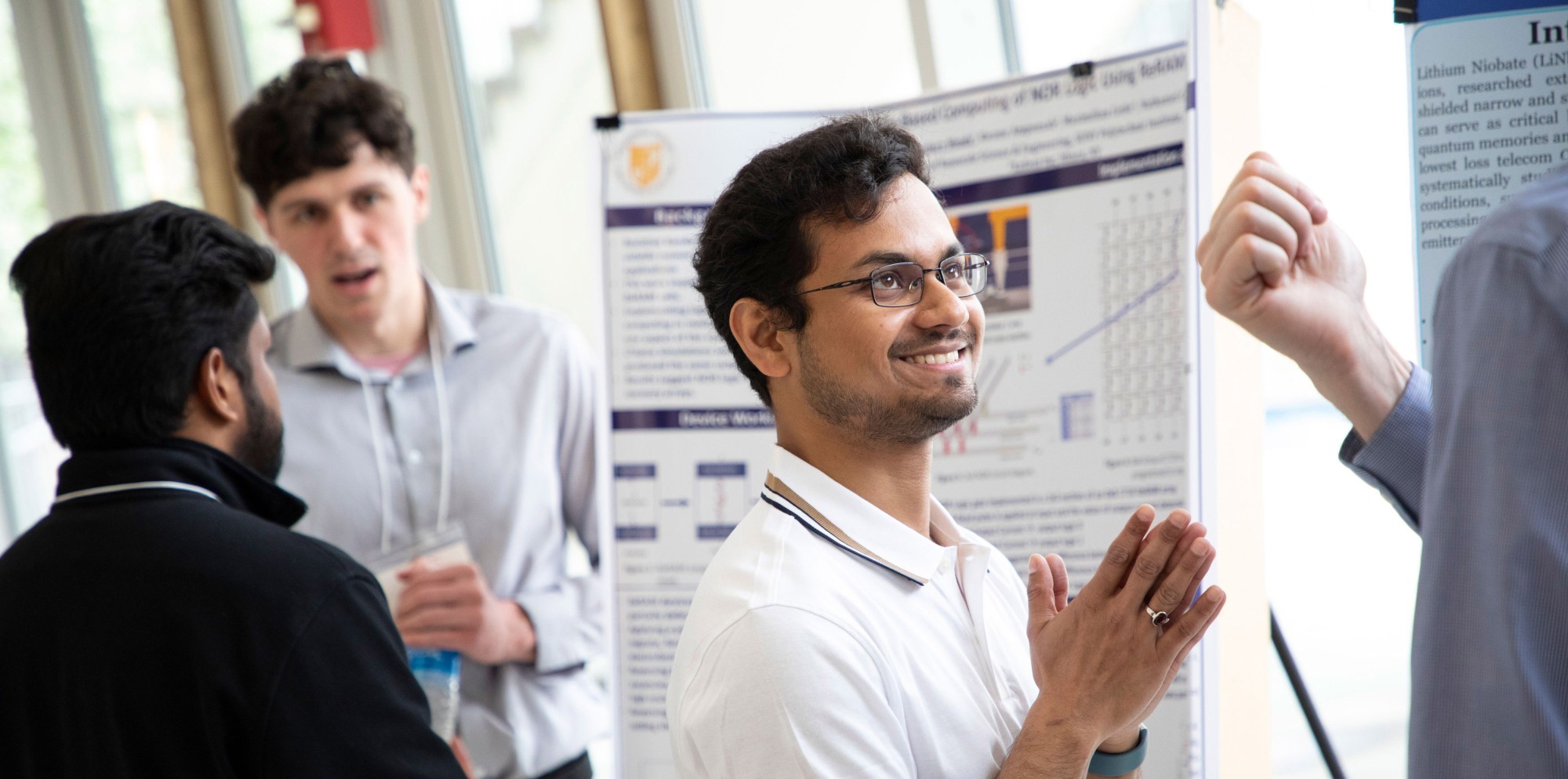 A student standing in front of a research poster smiles and puts his palms together as he listens to someone off camera at UAlbany Showcase.