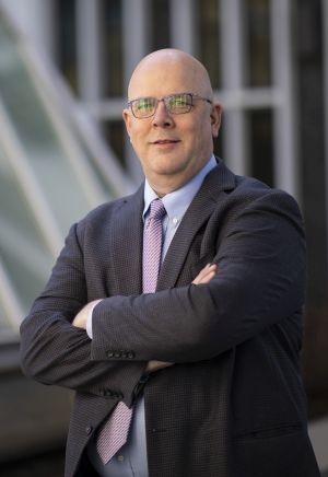 a man in glasses and a gray suit stands, arms crossed