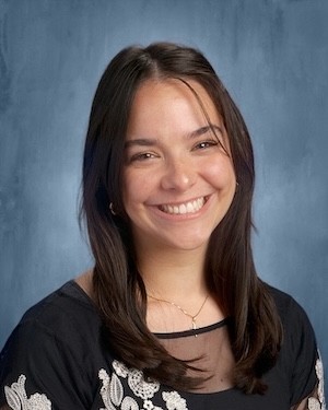 A young woman with long dark hair in a black blouse smiles for a portrait against a blue backdrop
