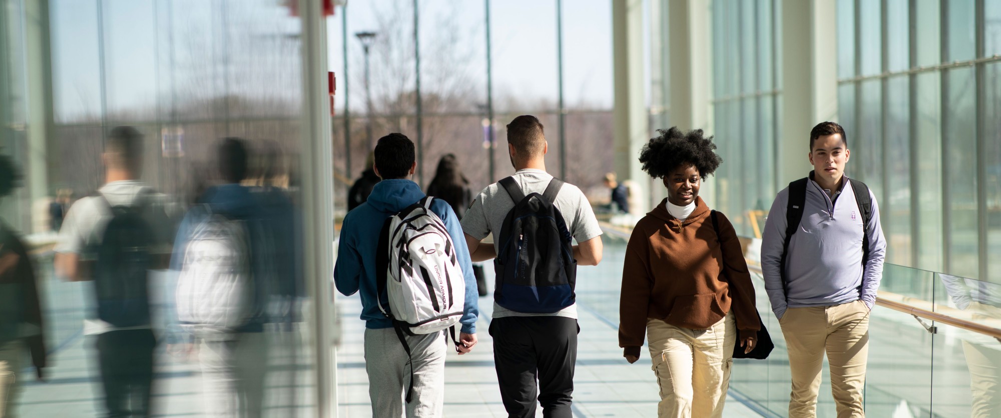 Four students with backpacks walk through the Massry Center for Business, which is a glass modern building on UAlbany's Uptown Campus.