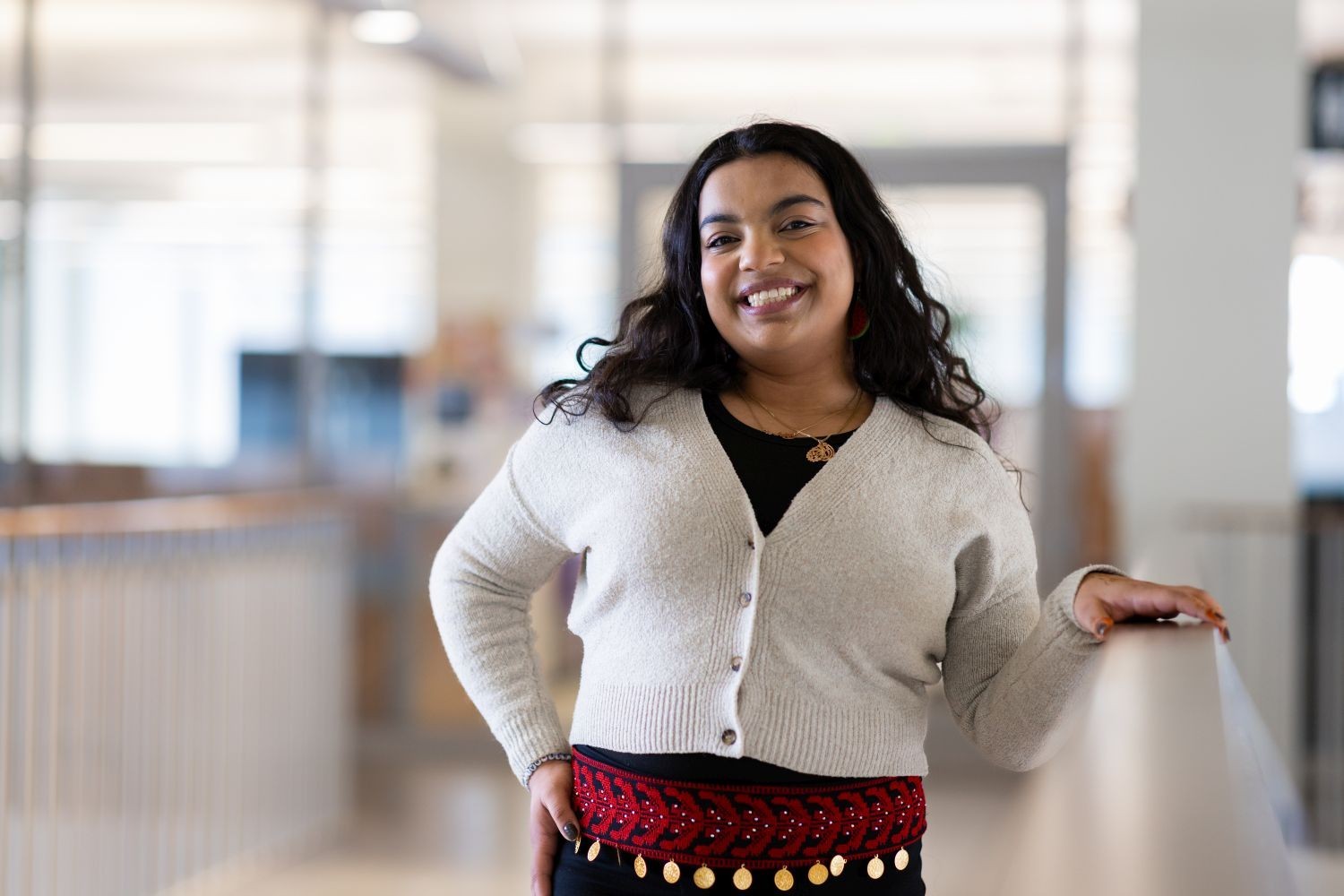 A smiling woman in a white sweater and a rad patterned sash worn as a belt