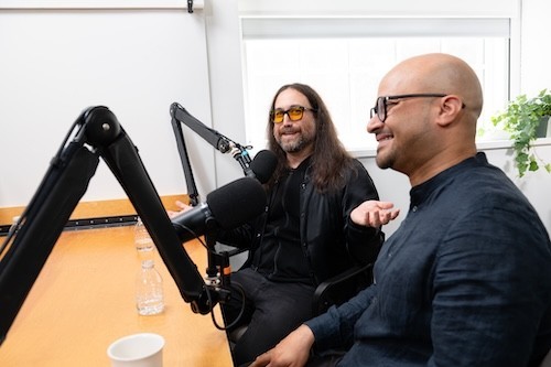 A man with long hair and glasses and a man with no hair and glasses smile and talk into microphones