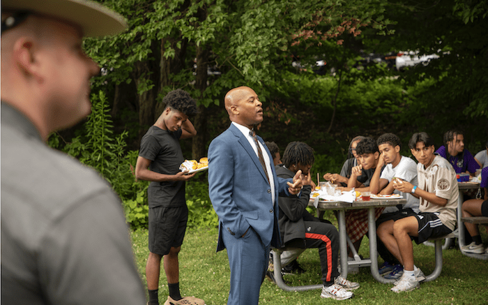 A man in a blue suit speaks to a crowd of students who are seated and standing at an outdoor pavilion in the woods. A state police trooper observes the scene in the foreground.