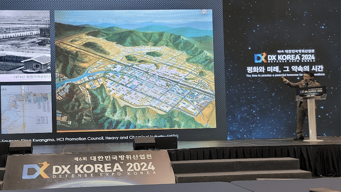 A man behind a podium gestures at a large screen showing a map and the title, DX Korea 2024.