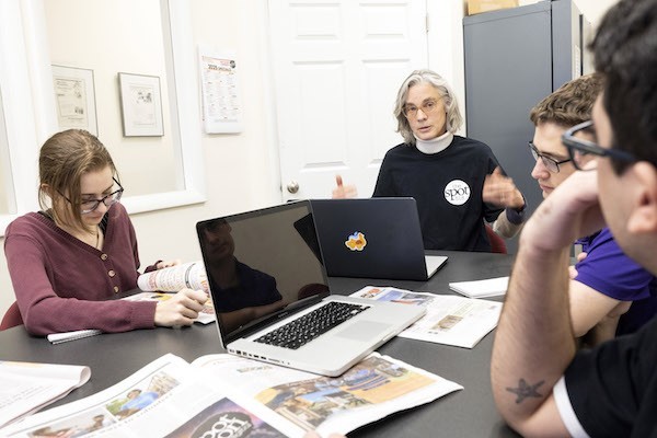 A woman with short gray hair talks to students seated at a table topped with laptops and newspapers.