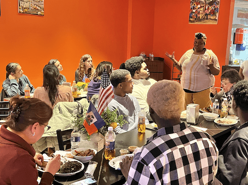 Students are seated around plates of food at a restaurant and listen as a woman in the front of the room speaks to the group.