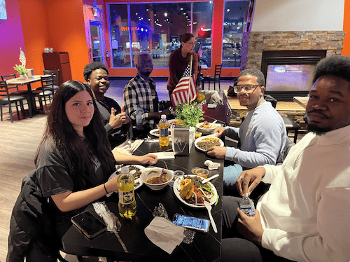 Students are seated at a table in a restaurant topped with plates of Haitian food and pose for a photograph