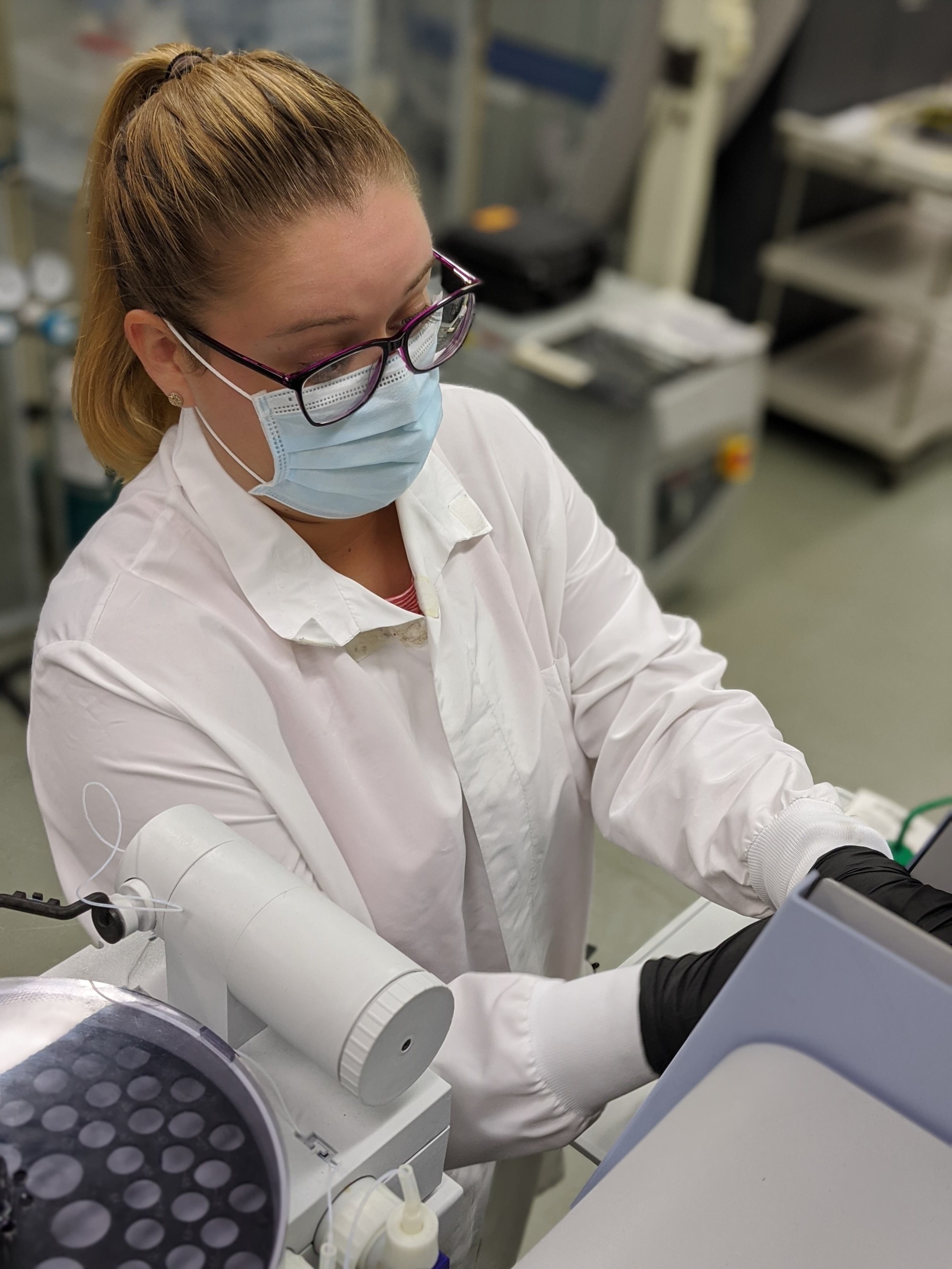 Emily wears a lab coat and a mask and sits at a desk in the Wadsworth Center.