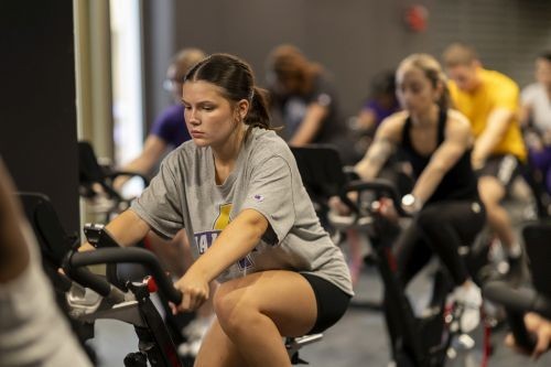 A woman rides a stationary bike in a gym; other cyclists are seen in the background