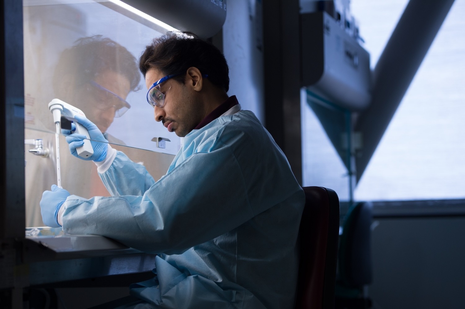 Man in blue research coat and protective goggles holding small white scientific equipment in a lab setting
