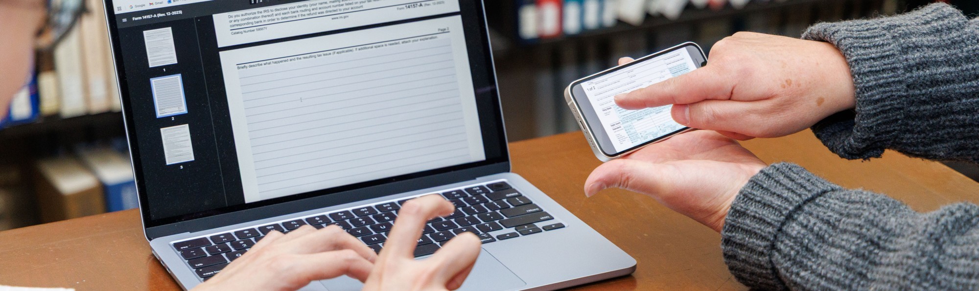 One person types on a laptop while a second person points to something on a smartphone's screen.