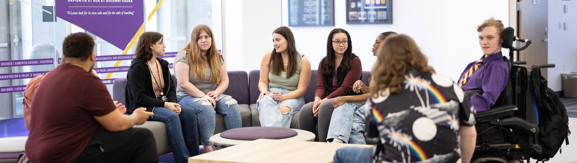 Eight UAlbany students sit in a circle in an academic building's lobby, chatting amongst themselves.