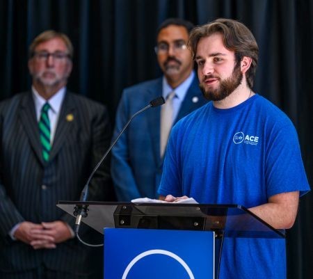 A young man speaks at a lecturn, with two others standing behind him 