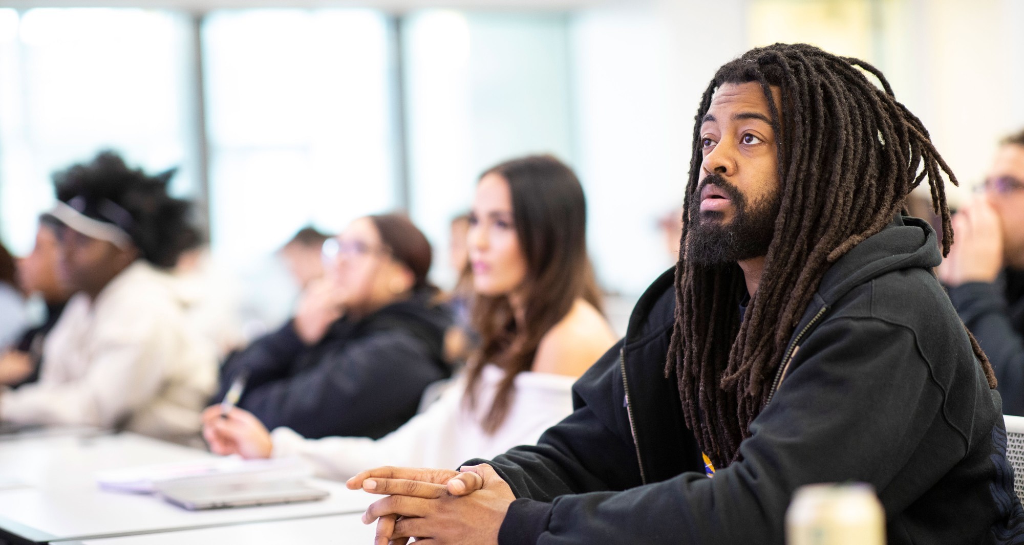 A student focused on a lecture sits at a desk in a classroom, with other students seated around him.