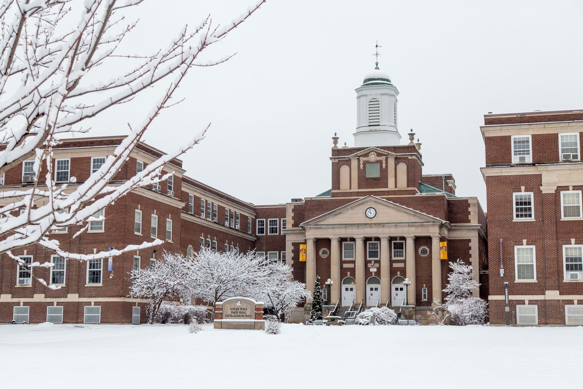 Brick buildings with UAlbany banners, surrounded by a snowy lawn.