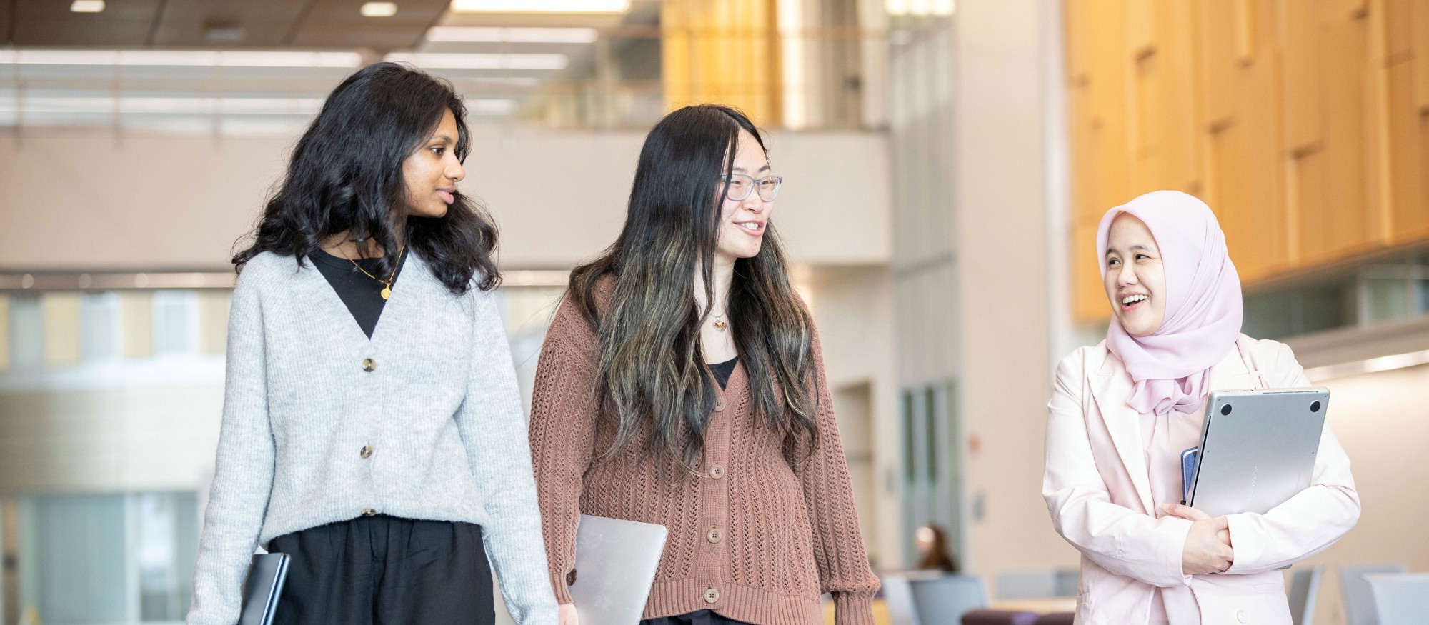 Three graduate students smile and chat as they walk through UAlbany's ETEC building carrying their laptops.