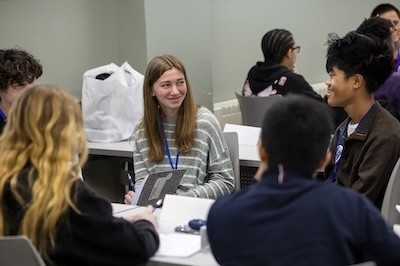 Teenage students gather around a table and smile as they chat and write in notebooks.
