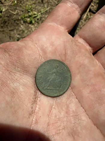 A dirt-covered hand holds an 18th century King George half penny in its palm with the ground in the background.
