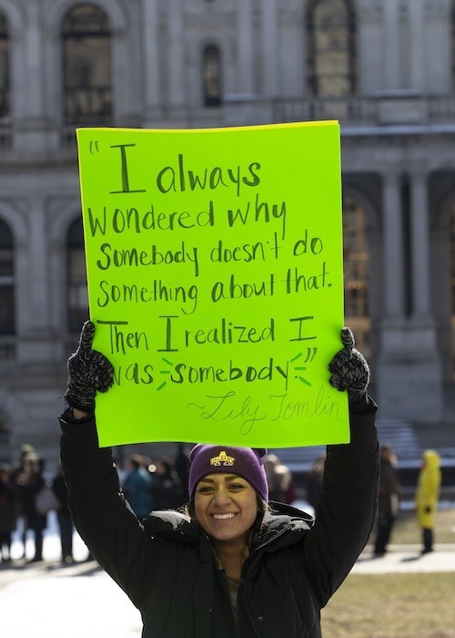 Student holds placard with quote from Lily Tomlin that reads "I always wondered why somebody doesn't do something about that. Then I realized I was somebody."