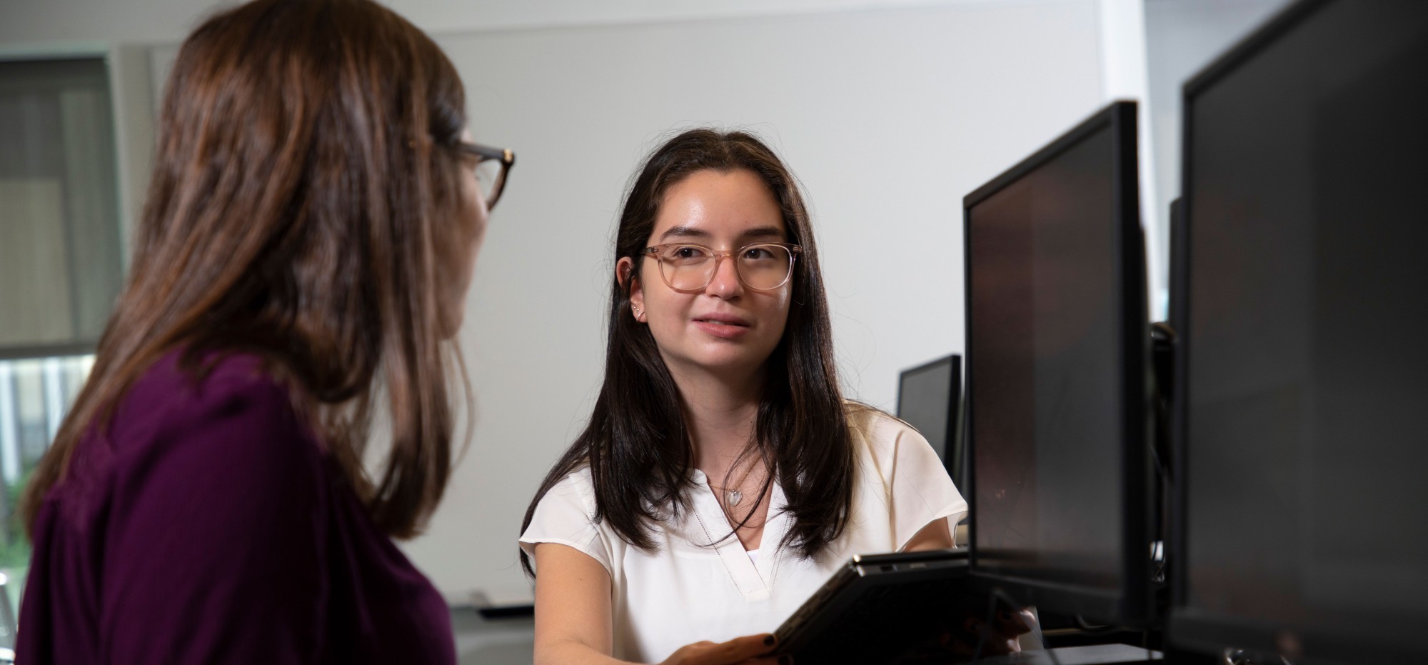 A student who is facing the camera and holding a clipboard looks at another student who is faced away from the camera and sitting at a desktop computer.
