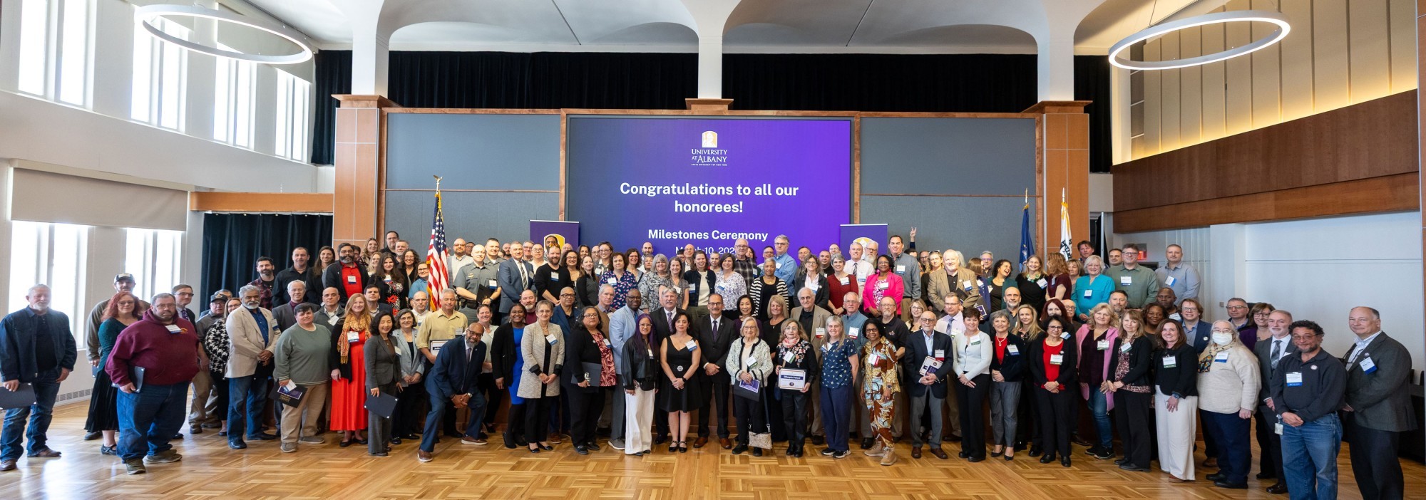 Over 100 people pose for a photo in front of a projector screen displaying the message, "Congratulations to all our honorees! Milestones Ceremony."