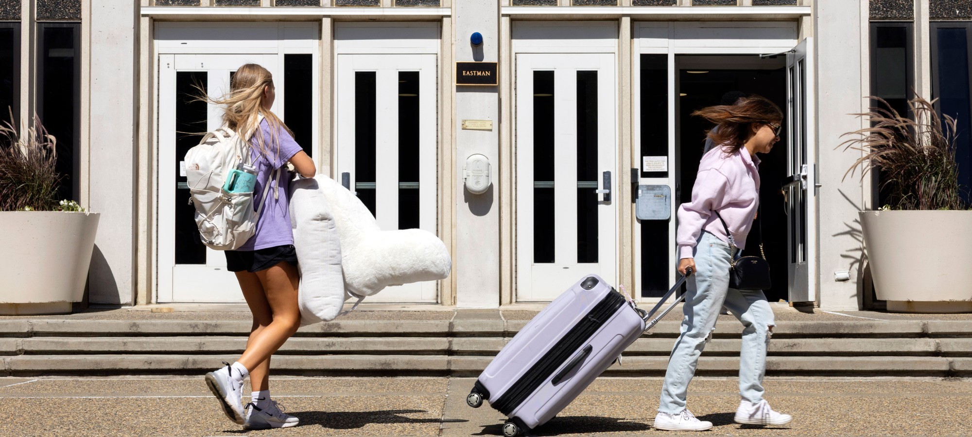 Two students carry belongings and roll suitcases into their residential hall on move-in day.