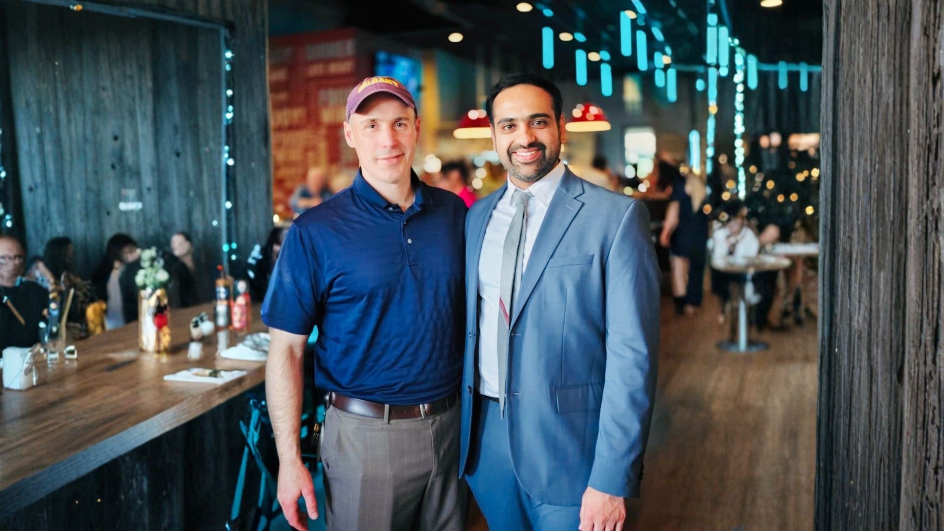 A smiling Nate Cady in a baseball hat and polo shirt stands next to a smiling Rajas Mathkari in a suit. a smiling Rajas 