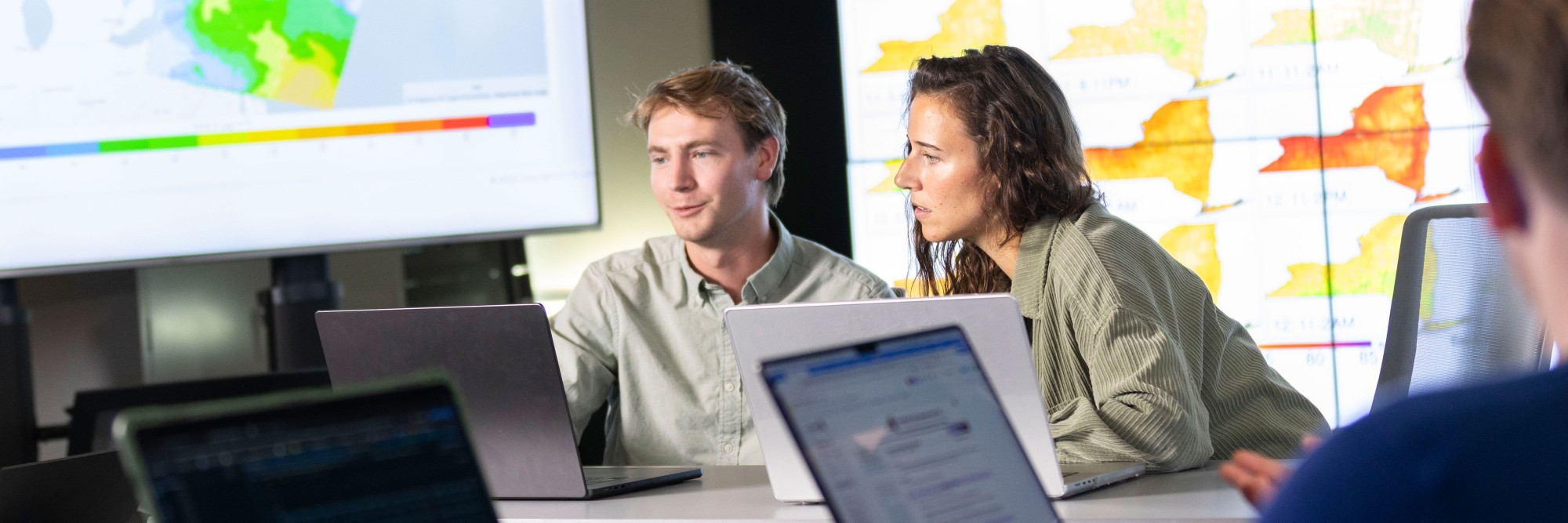 Two researchers work on laptops inside a lab space where maps are projected on screens around them.