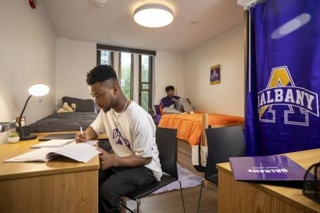 A student writes in a notebook at a desk in a dorm room; in the background another student is seen sitting on a bed.