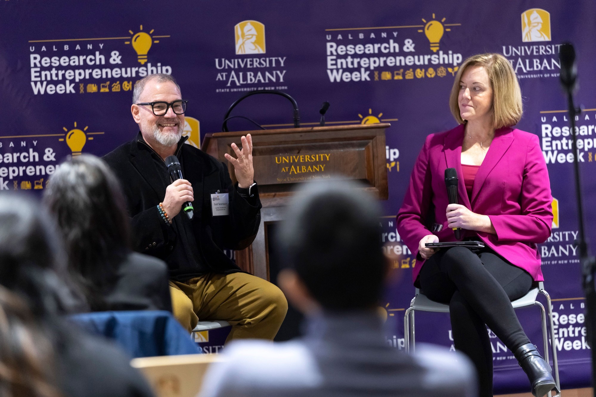 A moderator looks on an smiles as a panelist speaks into a handheld microphone at R&E Week 2025.