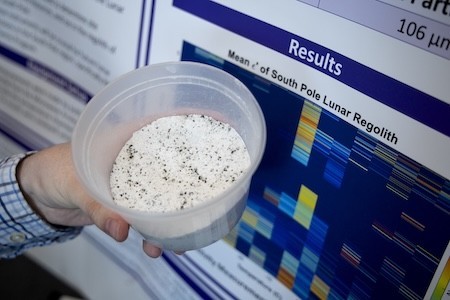 A hand holds a cup of lunar regolith simulant in front of a research poster