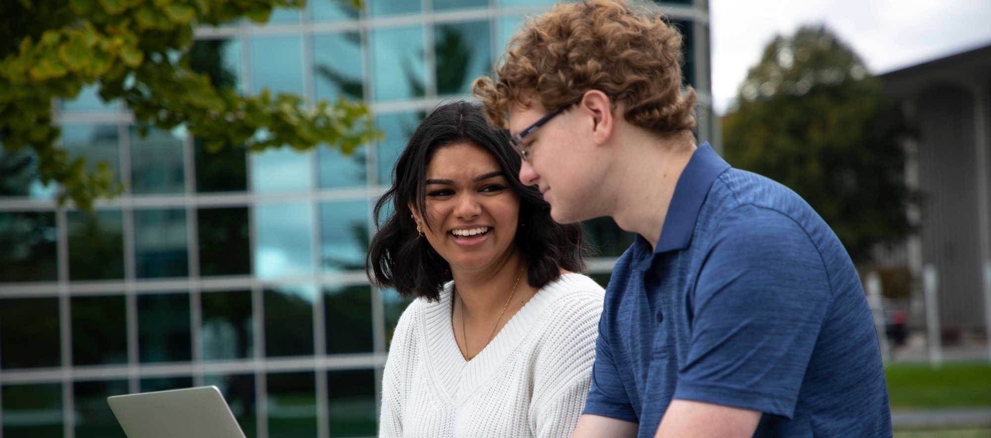Two students sit on an outdoor bench and smile as they collaborate on a laptop.