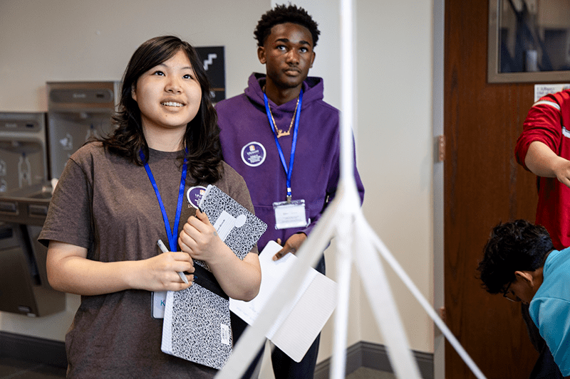 A teen girl holding a composition notebook and a teen boy in a purple hoodie look at a freestanding tower made out of paper and masking tape.