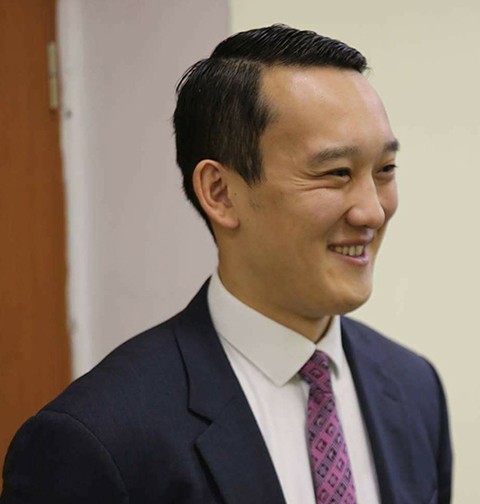 A man in a dark suit and purple tie stands in front of a brown and white background