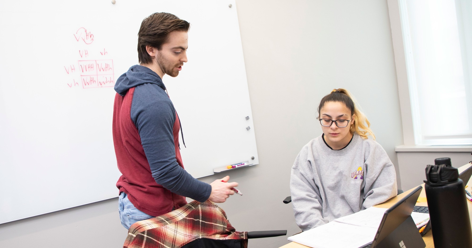 A tutor stands next to a white board and looks down at a computer in front of a seated student inside a tutoring room.