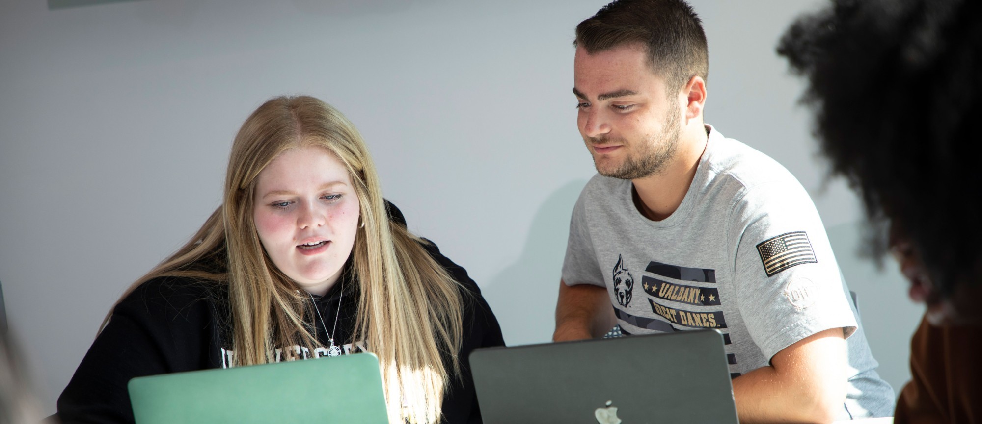 Two students sit together at a table with laptops open in front of them. One student smiles and looks on as the other student gestures and talks.