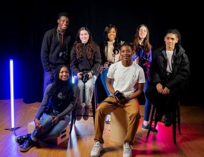 Seven students pose, seated in a group, with cameras in their laps or strapped around their necks