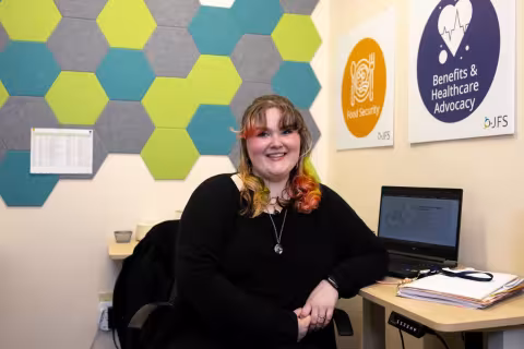 A young woman with rainbow hair smiles for a portrait while seated at a desk. Signs on the wall say "food security" and "benefits & advocacy".