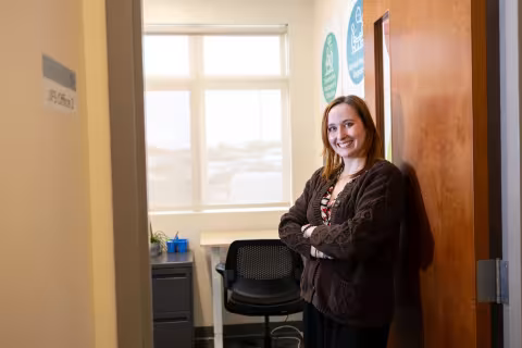 A young woman wearing a brown sweater smiles, arms folded, in the doorway to a warmly lit office