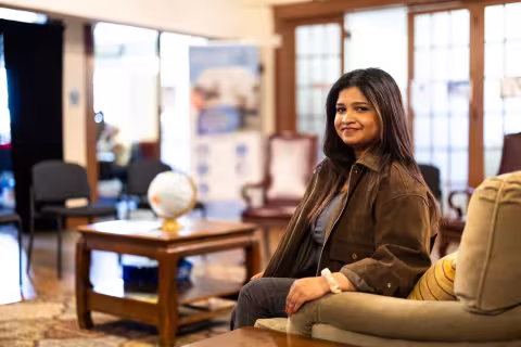 A woman poses for a portrait in an armchair. A globe sits on a coffee table behind her, with a bank of glass windows in the background.  