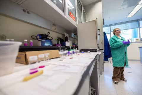 A woman wearing a green lab coat stands beside a lab bench containing an assortment of clear vials containing a purple substance.